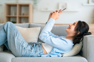 Mutlu, pozitif, genç, Asyalı bir kadın evde telefonda, gülümseyen bir kız. Uygulama kullanıyor ve internette akıllı telefonuyla kanepeye uzanıyor.