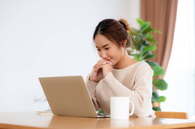 Oturma odasında dizüstü bilgisayarla çalışan mutlu genç Asyalı kadın. Serbest çalışan kadın masa başında oturmuş Webinar izlerken notlar yazıyor, internetten ders çalışıyor.
