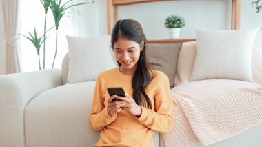Happy young asian woman relax on comfortable couch at home texting messaging on smartphone, smiling girl use cellphone, chatting online message, shopping online from home