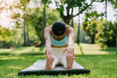 Genç Asyalı kadın, güneşli bir günde arka planda ağaçlarla yeşil çimlerde yoga yapıyor. Fitness, egzersiz ve açık hava egzersizi kavramı