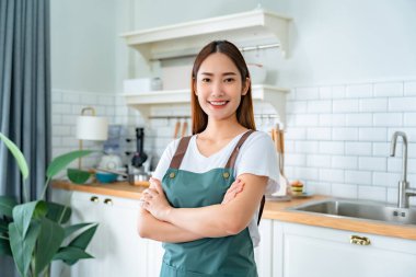 Önlük giyip mutfak odasında duran mutlu Asyalı kadın. Güzel bayan gülümsüyor ve kameraya bakıyor. Kendinden emin hissediyor.