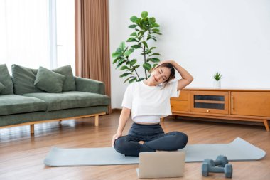 Genç Asyalı kadın oturma odasında bilgisayarlı dizüstü bilgisayarla antrenman yaparken yoga antrenmanını spor minderinde yapıyor.