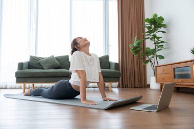 Genç Asyalı kadın oturma odasında bilgisayarlı dizüstü bilgisayarla antrenman yaparken yoga antrenmanını spor minderinde yapıyor.