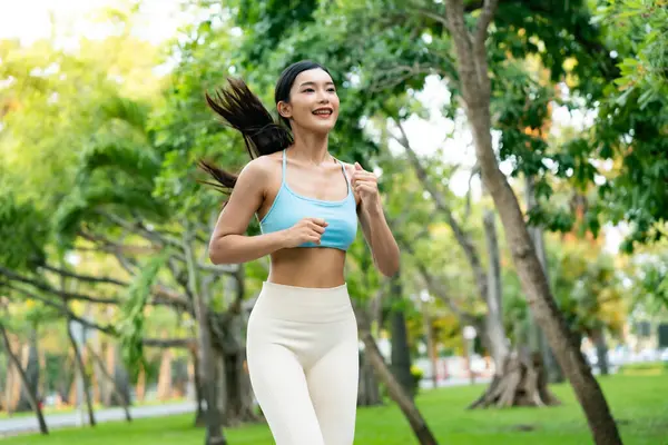 Asyalı bir kadın halka açık bir parkta koşuyor, atletik kıyafetler giyiyor ve yeşil, doğal bir ortamda kardiyo egzersizinin tadını çıkarıyor.