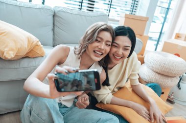 İki genç Asyalı kadın gülümseyip mutlu olurken evlerindeki oturma odalarında fotoğraf çektirip kameraya bakıyorlar.