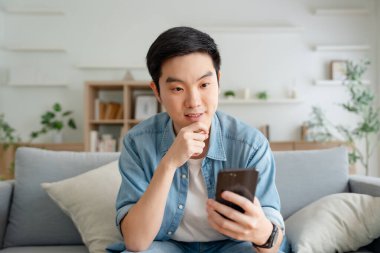 Oturma odasında komik mobil uygulamalar kullanarak rahat koltukta oturan gülümseyen yakışıklı Asyalı genç adam. Evdeki adam, online alışveriş yapıyor, arkadaşlarına mesaj atıyor, sosyal medyada paylaşıyor.