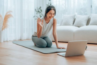 Genç Asyalı bir kadın laptopun önünde yoga minderinin üzerinde yerde yatıyor ve evde antrenman yapıyor. Egzersiz yapmadan önce internetten antrenörle iletişim kuran güzel, gülümseyen bir kadın.