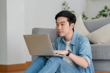 Genç Asyalı adam rahat koltukta dinleniyor, evde dizüstü bilgisayar kullanıyor, sosyal ağda arkadaşlarıyla sohbet ediyor, haftasonunu geçiriyor, film izliyor, online alışveriş yapıyor.