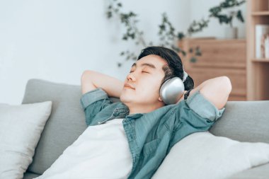 Rahat genç Asyalı adam rahat koltukta dinleniyor, sakin çekici adam kanepede dinleniyor, temiz hava soluyor gözleri kapalı, kulaklık takıyor.