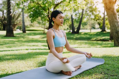 Barışçıl bir park ortamında meditasyon yapan Asyalı kadın, güneşli bir günde arka planda ağaçlarla yeşil çimlerde bacak bacak üstüne atarak oturuyor. Farkındalık, kişisel bakım ve açık hava rahatlığı kavramı