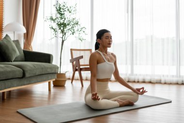 Genç Asyalı kadın evde nilüfer pozisyonunda meditasyon yapıyor, spor kıyafetleriyle yerde oturuyor. Rahatlama, huzur ve farkındalık için nefes alma egzersizi.