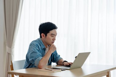 Oturma odasında dizüstü bilgisayarla çalışan mutlu Asyalı genç adam. Serbest çalışan erkek, masa başında oturmuş Webinar izlerken notlar yazıyor, internetten çalışıyor.