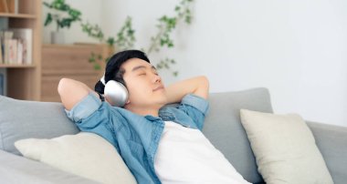 Rahat genç Asyalı adam rahat koltukta dinleniyor, sakin çekici adam kanepede dinleniyor, temiz hava soluyor gözleri kapalı, kulaklık takıyor.