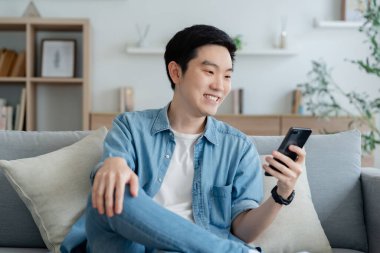 Oturma odasında komik mobil uygulamalar kullanarak rahat koltukta oturan gülümseyen yakışıklı Asyalı genç adam. Evdeki adam, online alışveriş yapıyor, arkadaşlarına mesaj atıyor, sosyal medyada paylaşıyor.