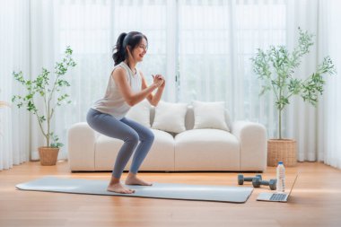 Genç Asyalı güzel kadın evde aerobik egzersizi yapıyor. Çekici kız çömelme egzersizi yapıyor, evdeki sağlık için çevrimiçi eğitmenin talimatlarını takip ediyor.