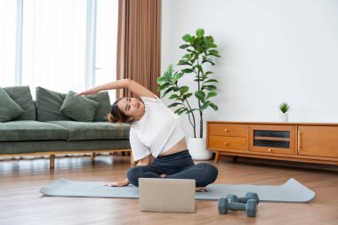 Genç Asyalı kadın oturma odasında bilgisayarlı dizüstü bilgisayarla antrenman yaparken yoga antrenmanını spor minderinde yapıyor.