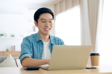 Oturma odasında dizüstü bilgisayarla çalışan mutlu Asyalı genç adam. Serbest çalışan erkek, masa başında oturmuş Webinar izlerken notlar yazıyor, internetten çalışıyor.