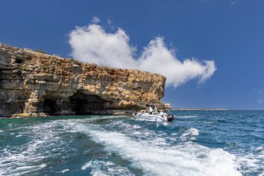 Polignano a Mare, İtalya, Apulia - 24 Mayıs 2024: Adriyatik Denizi kıyısındaki pitoresk uçurumların suyundan görüş. Turistler kayalıklardaki bir mağaraya tekne ile giderler.