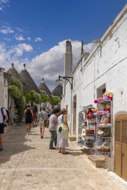Alberobello, Apulia, İtalya - 21 Mayıs 2024: Trulli evleri, geleneksel, eşsiz, konik çatılı Apulian kuru taş kulübesi. Caddelerden birinde hediyelik eşya dükkanlarıyla yürüyen turistler.
