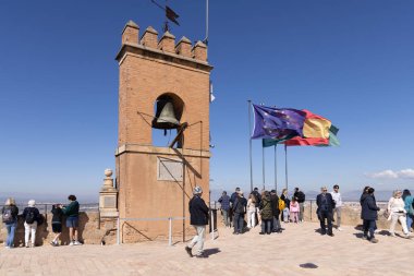 Granada, İspanya, Endülüs - 23 Mart 2025: Alhambra Kalesi 'nin Gözcü Kulesi' nin (Torre de la Vela) üzerindeki tarihi çan ve bayraklar. Ünlü anıtı ziyaret eden bir grup turist