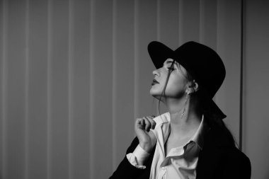 Stüdyoda poz veren siyah ceketli ve şapkalı güzel bir genç kızın siyah beyaz fotoğrafı.