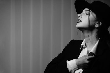 Stüdyoda poz veren siyah ceketli ve şapkalı güzel bir genç kızın siyah beyaz fotoğrafı.