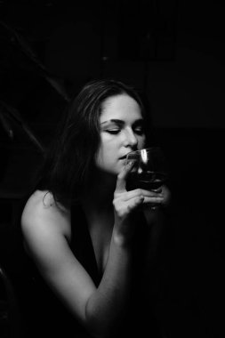 Siyah elbiseli güzel bir kadının siyah beyaz fotoğrafı. Bir bardak viski ile kameraya poz veriyor.