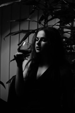 Siyah elbiseli güzel bir kadının siyah beyaz fotoğrafı. Bir bardak viski ile kameraya poz veriyor.