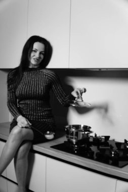 Çizgili kısa elbiseli hoş bir kadının siyah beyaz fotoğrafı mutfaktaki sobanın yanındaki masada oturuyor.