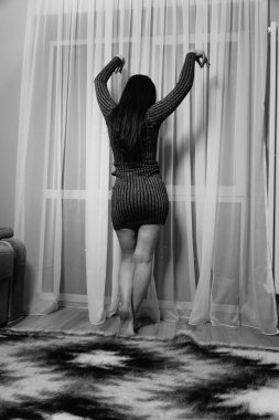 Siyah beyaz bir fotoğraf çizgili kısa elbiseli hoş bir kadın odada perdelerin yanında duruyor.