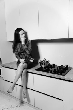 Çizgili kısa elbiseli hoş bir kadının siyah beyaz fotoğrafı mutfaktaki sobanın yanındaki masada oturuyor.