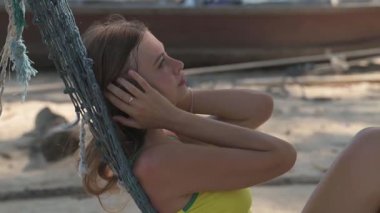 Sarı kolsuz tişörtlü sarışın kadın tropik bir plajda palmiye ağacının altında dinleniyor. Yatmakla dik oturmak arasında gidip geliyor. Arka planda palmiye ağaçları, kayalar ve tekneler var..