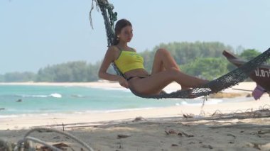 Sarı kolsuz tişörtlü sarışın kadın tropik bir plajda palmiye ağacının altında dinleniyor. Yatmakla dik oturmak arasında gidip geliyor. Arka planda palmiye ağaçları, kayalar ve tekneler var..