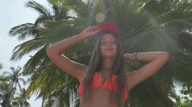 Sarışın kadın palmiye ağacının önünde duruyor, gülümsüyor ve ellerini sallıyor. Güneş ışığı yaprakların arasından süzülür, tropik plaj arka planında palmiye ağaçları, kayalar ve tekneler vardır..