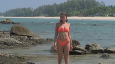 Turuncu bikinili güzel sarışın bir kadın tropik bir plajda yürüyor, gülümsüyor ve güneşli havanın tadını çıkarıyor. Arka planda palmiye ağaçları, kayalar ve geleneksel tekneler yer alıyor.