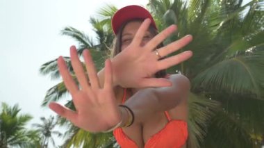 Sarışın kadın palmiye ağacının önünde duruyor, gülümsüyor ve ellerini sallıyor. Güneş ışığı yaprakların arasından süzülür, tropik plaj arka planında palmiye ağaçları, kayalar ve tekneler vardır..