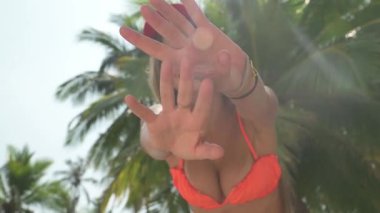 Sarışın kadın palmiye ağacının önünde duruyor, gülümsüyor ve ellerini sallıyor. Güneş ışığı yaprakların arasından süzülür, tropik plaj arka planında palmiye ağaçları, kayalar ve tekneler vardır..