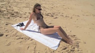 Kahverengi bikinili kadın kumsalda beyaz bir battaniyeye oturmuş, elinde şnorkel maskesi var. Arkasında, kayalık bir sahil ve tropikal palmiye ağaçları huzur dolu sahneyi tamamlıyor.