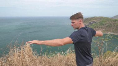 Dağda yoga yapan atletik bir adam uzak adaları olan sakin bir deniz manzarasına bakıyor. Tropikal, manzaralı bir ortamda sakin bir farkındalık anı.