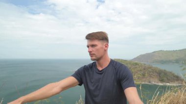 Dağda yoga yapan atletik bir adam uzak adaları olan sakin bir deniz manzarasına bakıyor. Tropikal, manzaralı bir ortamda sakin bir farkındalık anı.