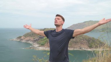 Dağda yoga yapan atletik bir adam uzak adaları olan sakin bir deniz manzarasına bakıyor. Tropikal, manzaralı bir ortamda sakin bir farkındalık anı.