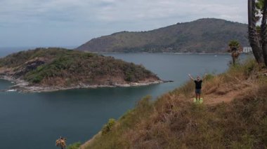 Uçurumun kenarında yoga yapan bir adam. İnsansız hava aracından çekilen çarpıcı okyanus ve ada manzaralı. Huzurlu, güçlü tropikal manzara.