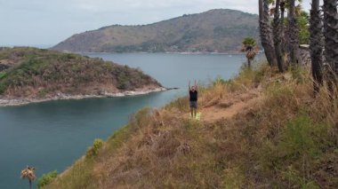 Uçurumun kenarında yoga yapan bir adam. İnsansız hava aracından çekilen çarpıcı okyanus ve ada manzaralı. Huzurlu, güçlü tropikal manzara.