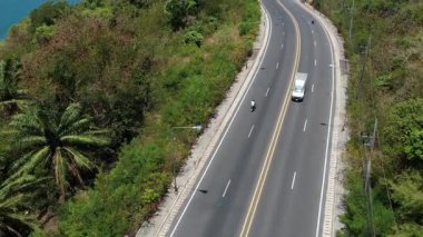 İnsanoğlu, okyanus manzaralı ve yakındaki adaları insansız hava aracı tarafından yakalanan dolambaçlı bir tropikal ada yolunda motosiklet sürüyor. Manzaralı ve maceralı.