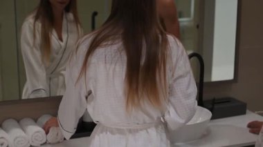 Çift aynanın karşısında duruyor, yüz kremi sürüyor, gülümsüyor ve sohbet ediyor. Sıcak, samimi bir cilt bakımı anı.