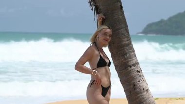 Siyah bikinili kadın, arka planda deniz olan palmiye yaprağı gölgelerinin altında duruyor..