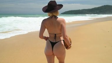 Şapkalı ve bikinili bir kız, arka planda turkuaz deniz ve uzak adalarla uzun bir tropikal sahilde yürüyor..