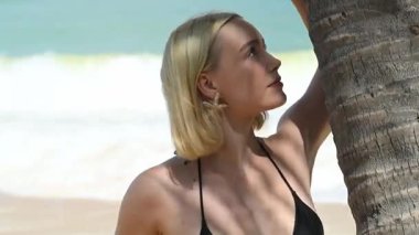 Gülümseyen kadın bir palmiye ağacının yanında durur, kameraya bakar okyanus ve arka planda nazik dalgalar.