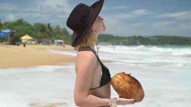 Arkasından köpüklü dalgalar düşerken tropik bir sahilde hindistan cevizi tutan genç bir kadın..