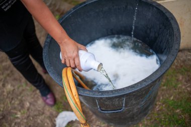Bir el, siyah kovaya sıvı sabun döküyor ve açık hava ortamında köpük oluşturuyor. Bu temizlik ve hijyen faaliyetlerinin özünü yakalar..